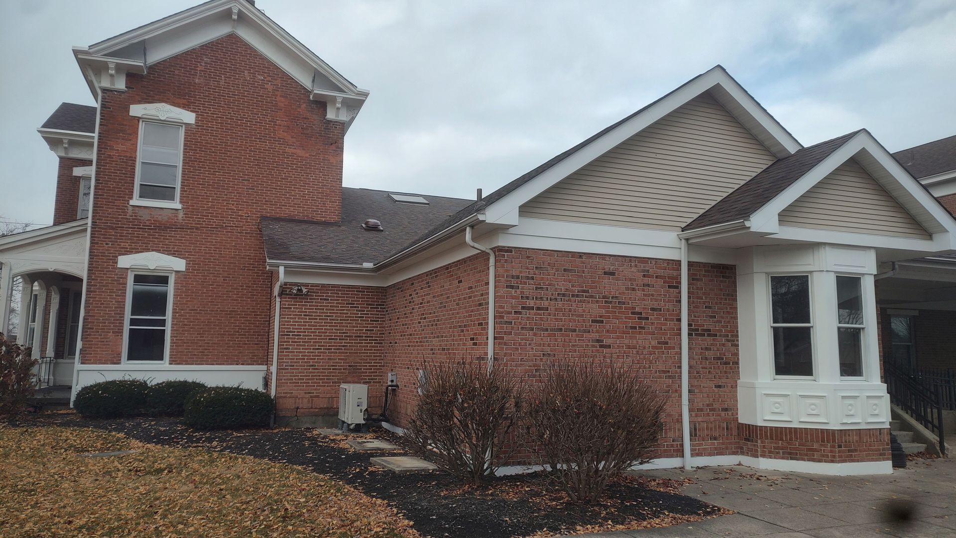 A red brick historic-style house with a side addition, a shingled roof, and a small bay window under a gray, cloudy sky.
