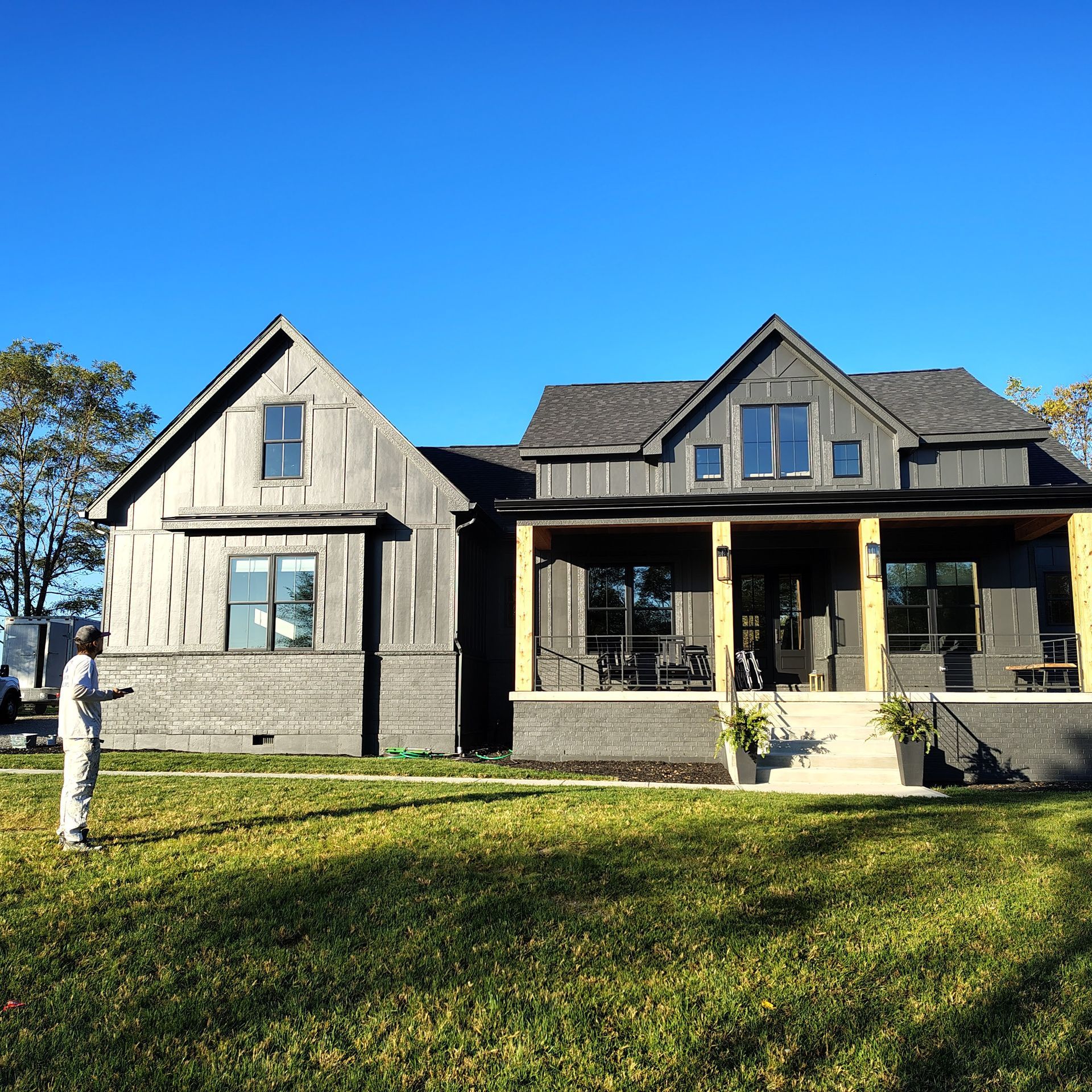 A person stands in a grassy yard before a newly built, dark gray farmhouse-style home with a covered front porch.