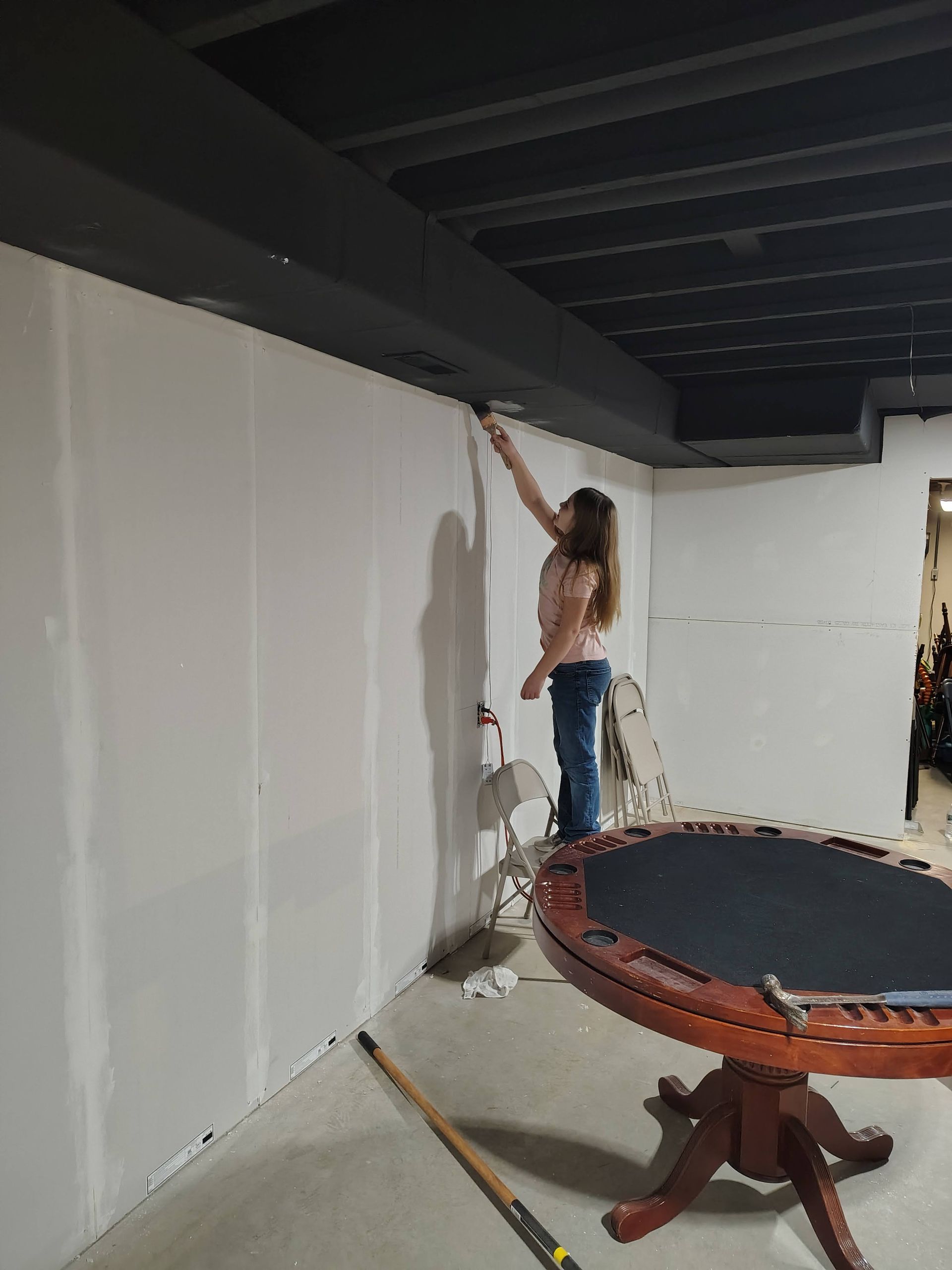 A person paints the trim along a black ceiling in an unfinished basement with a white wall and a wooden table.