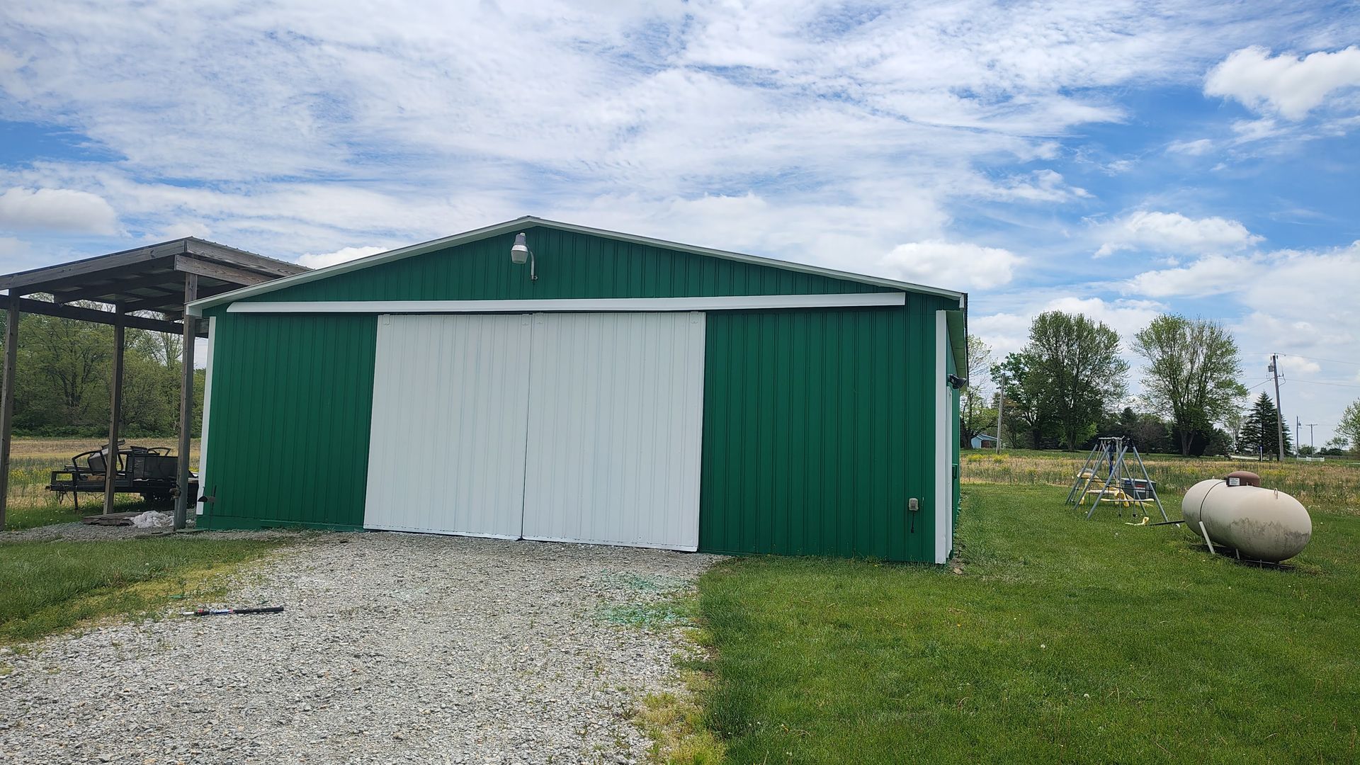 A green metal workshop with large white sliding doors sits on a gravel path in a rural, grassy field under a cloudy sky.