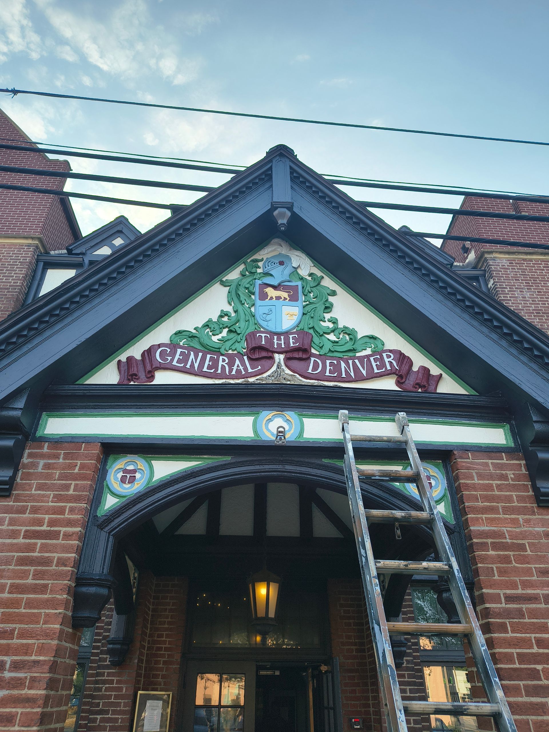 A ladder leans against the ornate brick entrance of The Golden Denver, a building with a dark-trimmed gable and crest.