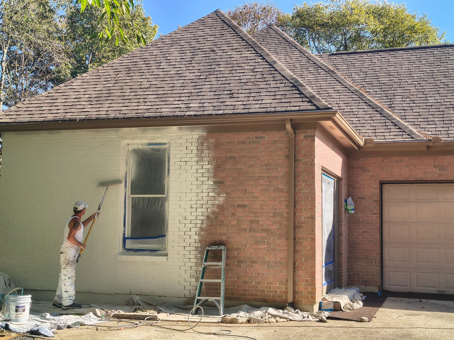 A person paints a brick house exterior from a warm red to a light cream color using a long-handled roller.