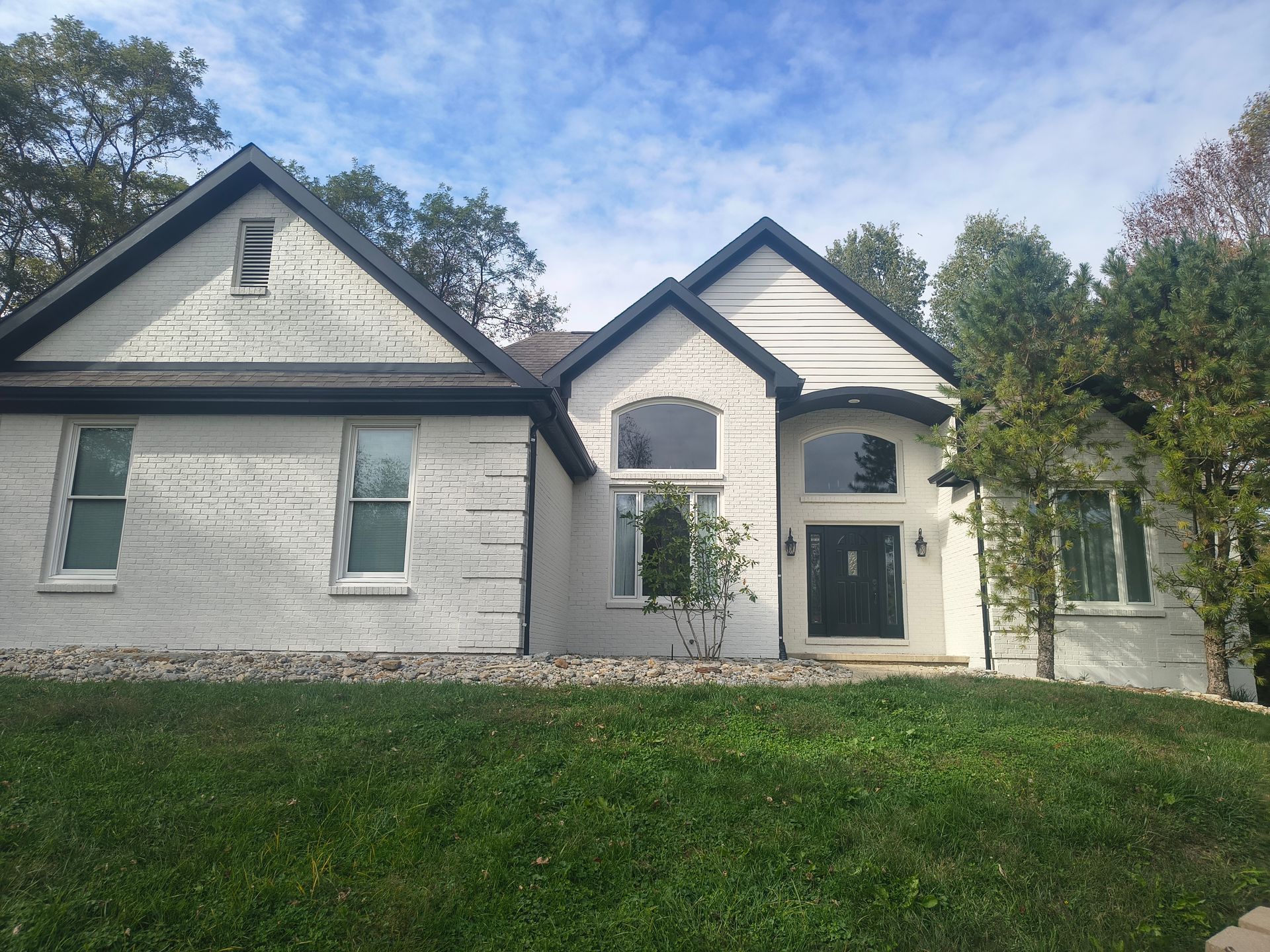 A two-story white house with dark trim and a gabled roof stands behind a sloping front lawn under a cloudy blue sky.