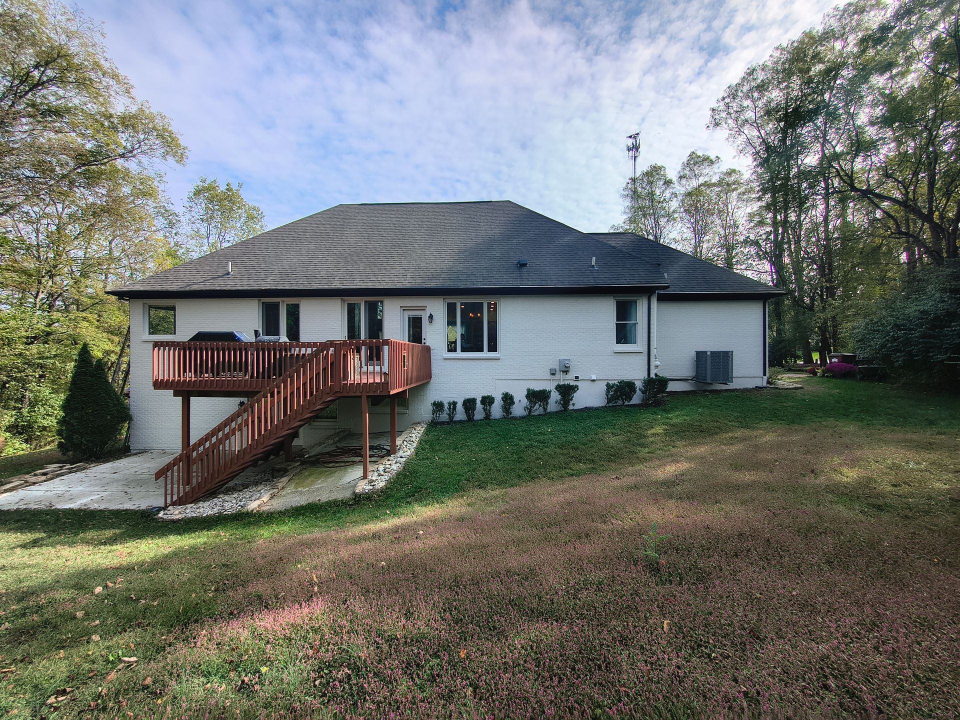A white, single-story house with a dark shingled roof, a wooden deck, and a staircase leading to a grassy backyard.