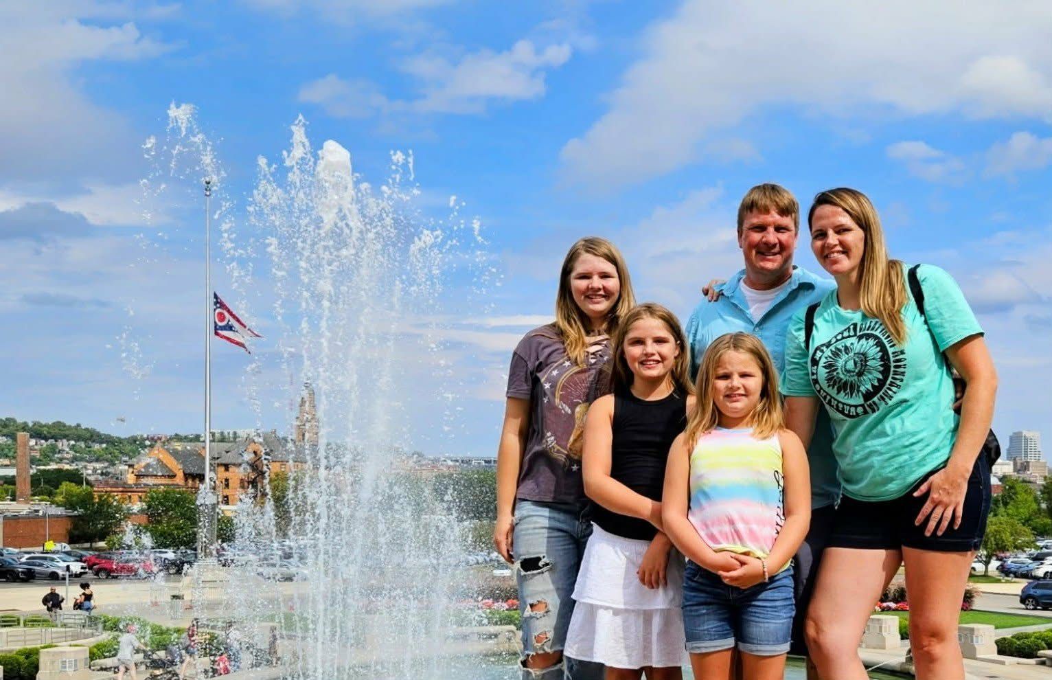 A family smiling for a photo in front of a fountain and a flag on a sunny day.