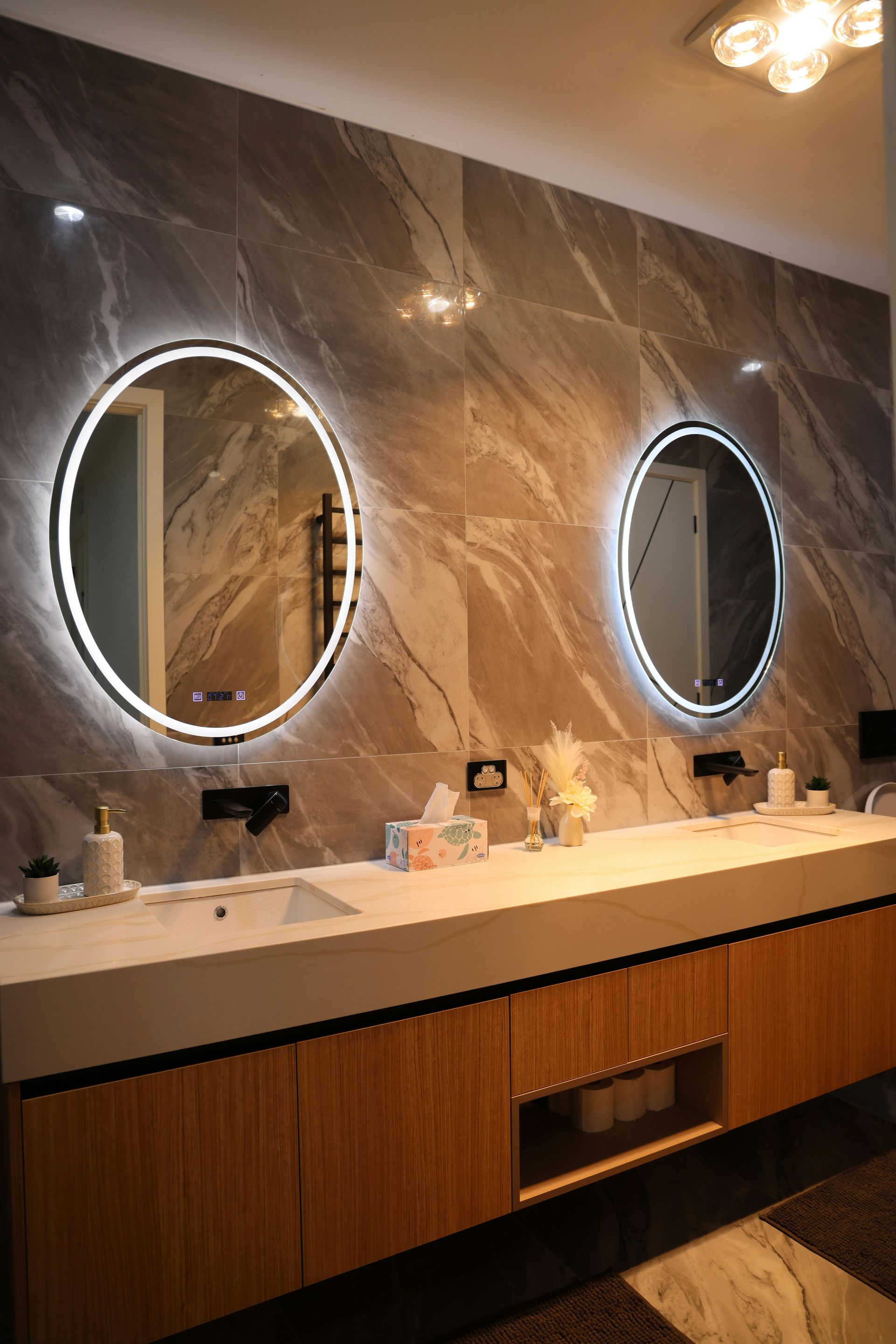 Two round mirrors with illuminated edges above a bathroom vanity with a marble accent wall.