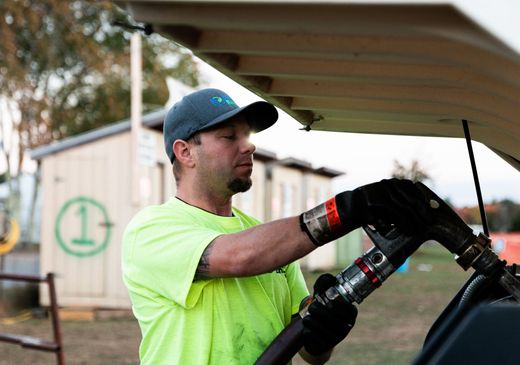 Man wearing a neon shirt and a cap fueling a vehicle outdoors near a small building.