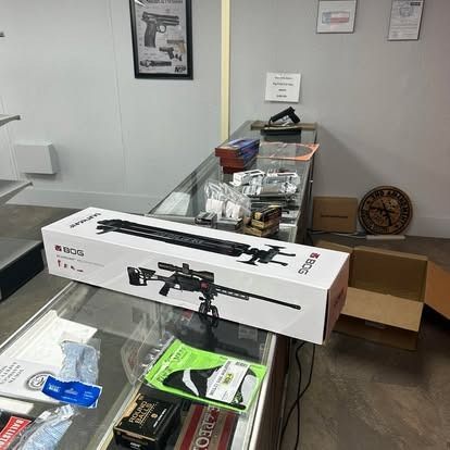 Inside a gun store, a rifle in its box sits on a glass counter next to other products and display items.