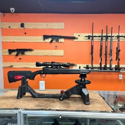 Rifle on display in a gun store, supported by a black stand, with other guns on the wall.