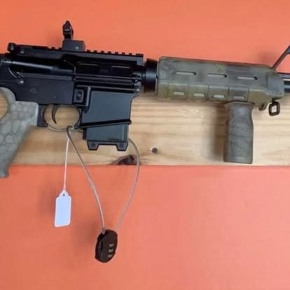 Black and tan rifle on a wooden surface, with a forward grip and security lock attached.