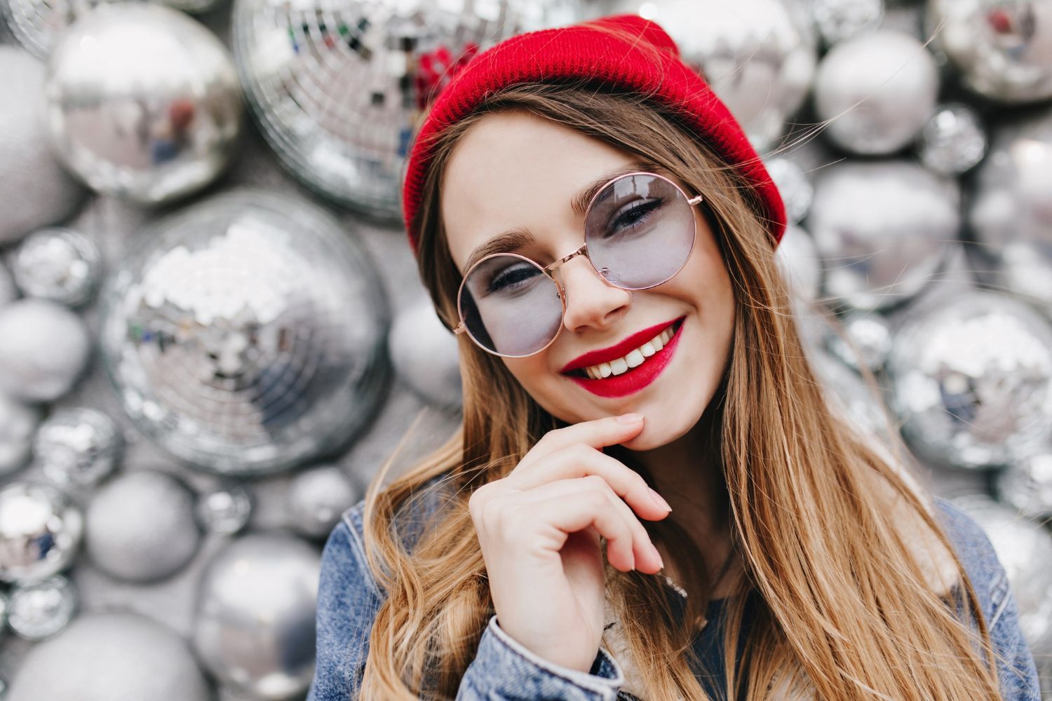Woman in red hat, sunglasses, and denim jacket smiling in front of silver orbs.