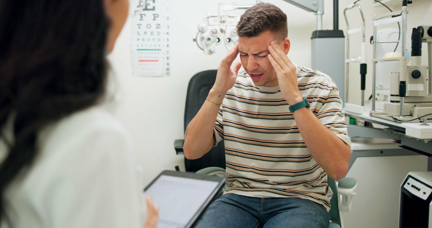 Man with a headache at the eye doctor's office, touching temples. Doctor in foreground.