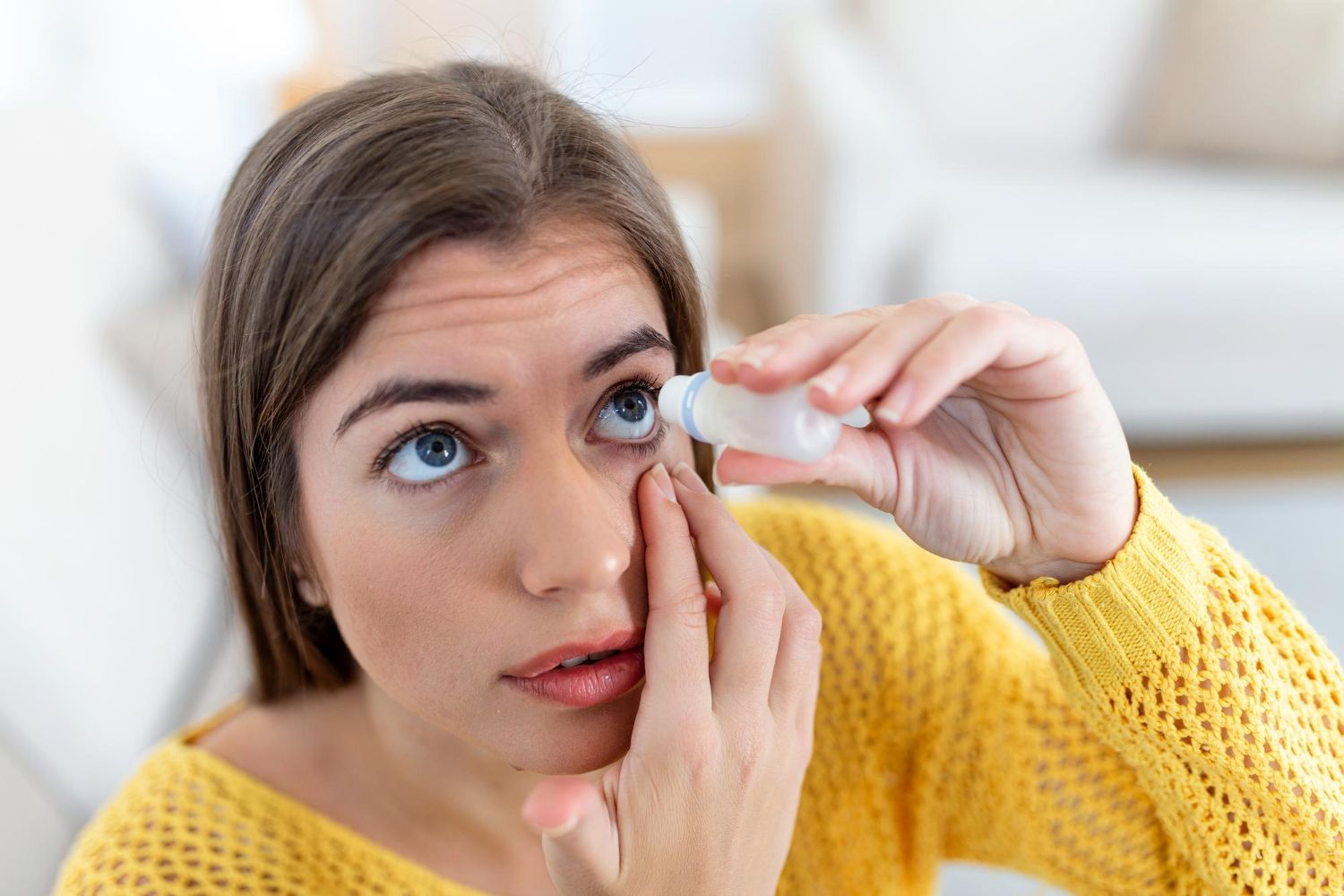 Woman in yellow sweater puts eye drops in her eye, looking upward, indoors.