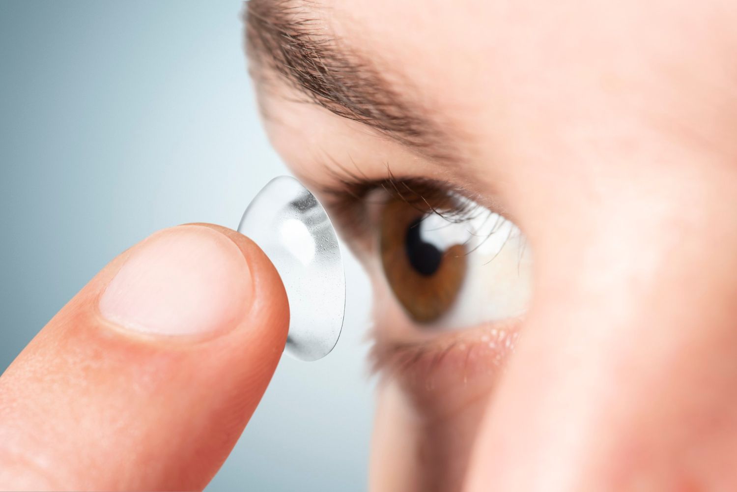 Close-up of a person putting a contact lens in their eye. Brown eye, light blue background.