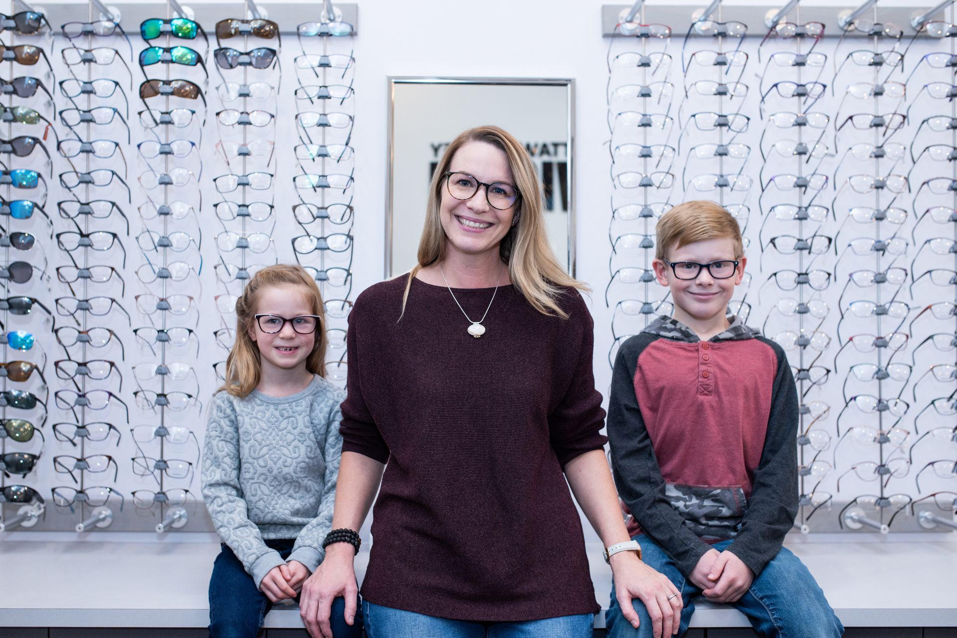 A woman and two children wearing glasses are sitting on a bench in front of a wall of glasses.