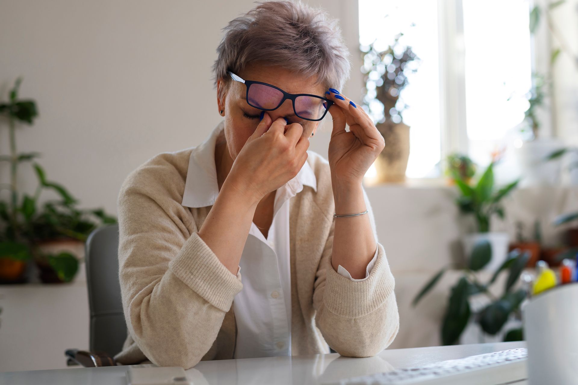 A woman wearing glasses is rubbing her eyes while sitting at a desk.