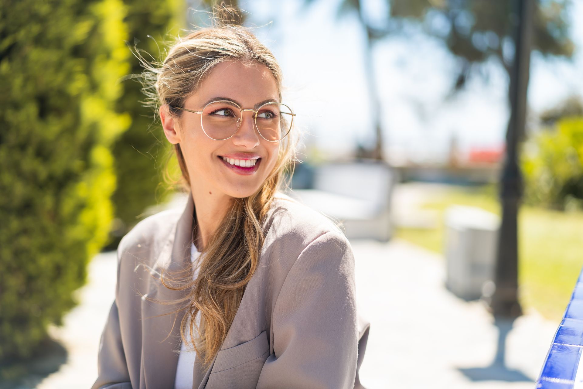 A woman wearing glasses is smiling while sitting on a bench in a park.