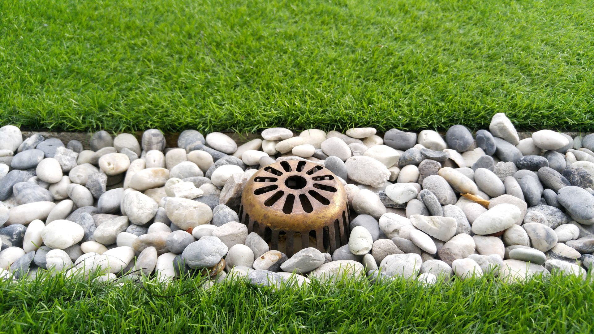 Drain in a bed of white and gray river rocks, between green grass borders.
