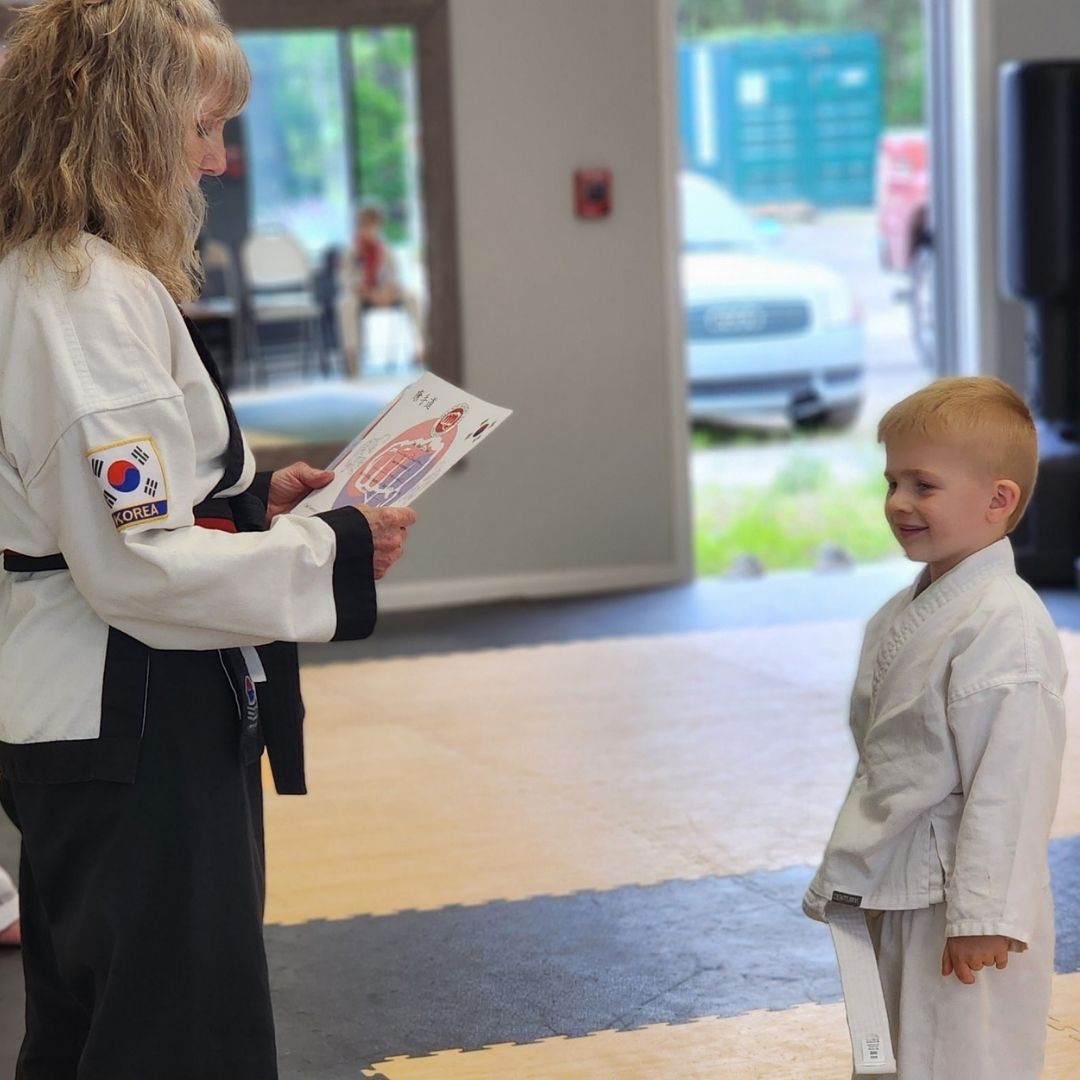 Two children practicing Brazilian Jiu-Jitsu on a mat. One child in white gi has the other in a leg lock. A girl watches.