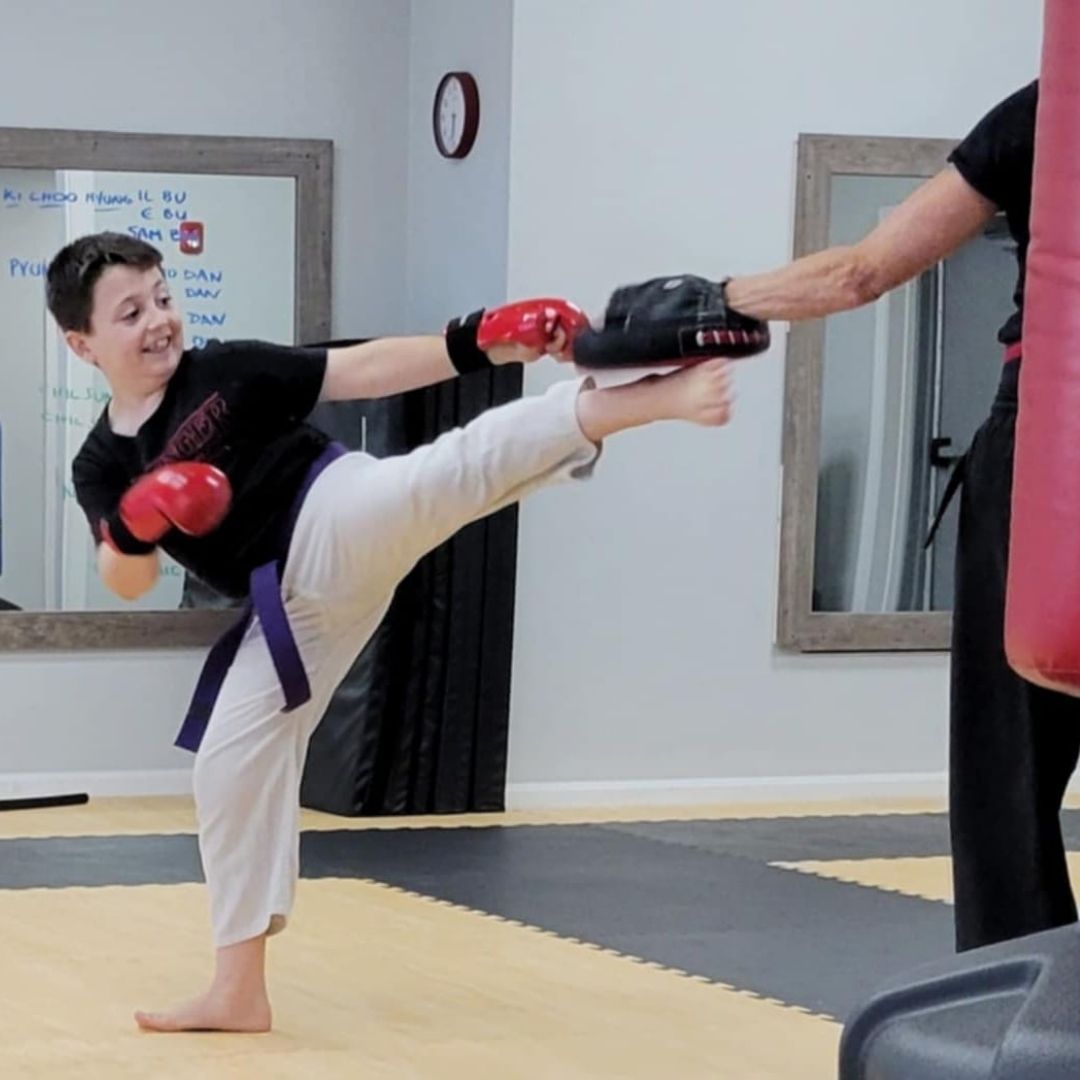 Two children in black and white martial arts uniforms practice a chokehold on a mat. One child smiles.