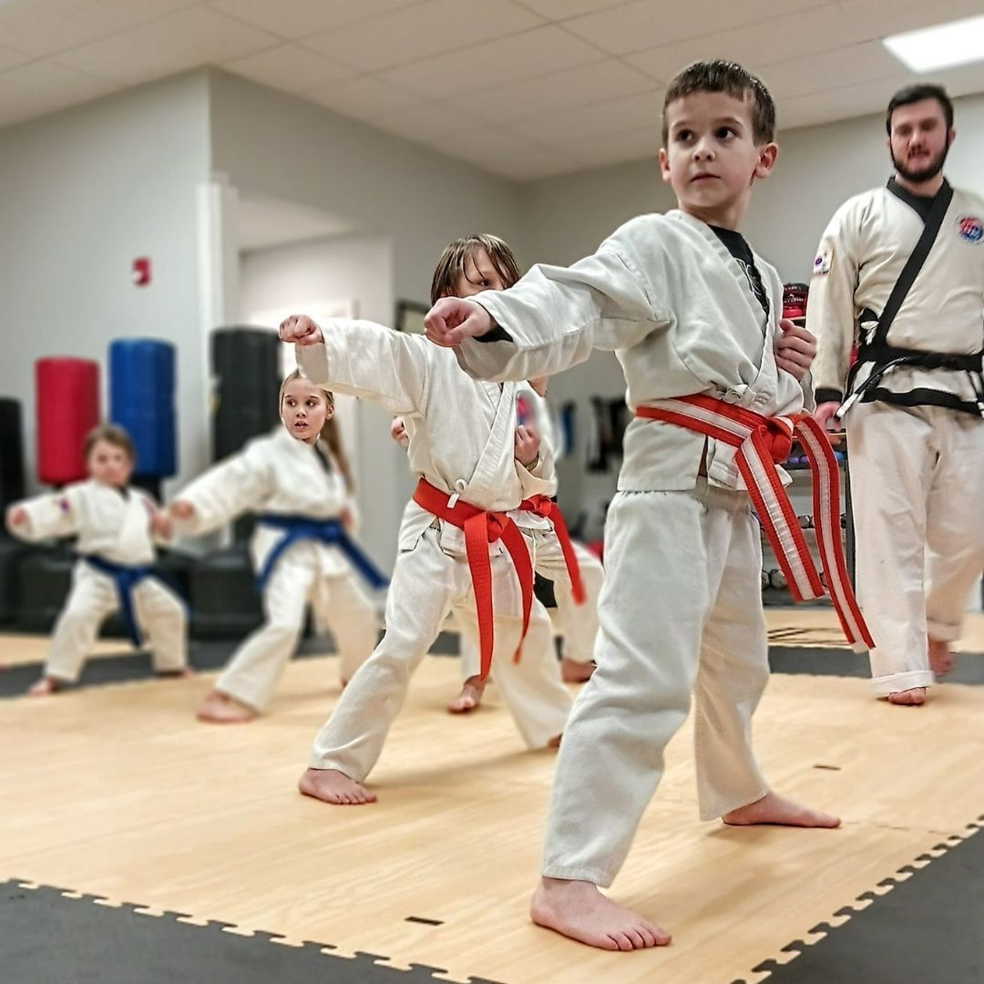 Children in colorful gis play a game on a mat. Two instructors pull a student in blue.