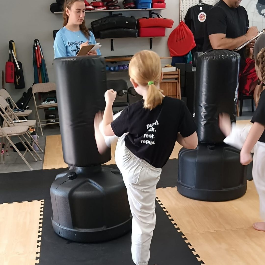 Two children practicing Brazilian Jiu-Jitsu on a mat. One child in white gi has the other in a leg lock. A girl watches.