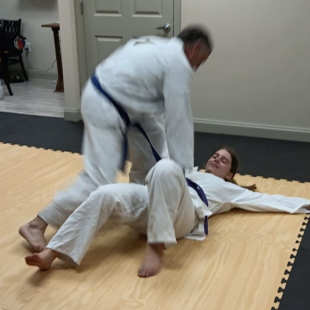 Two men in grappling position on a mat. One embraces the other from behind, both smiling. Gym setting.