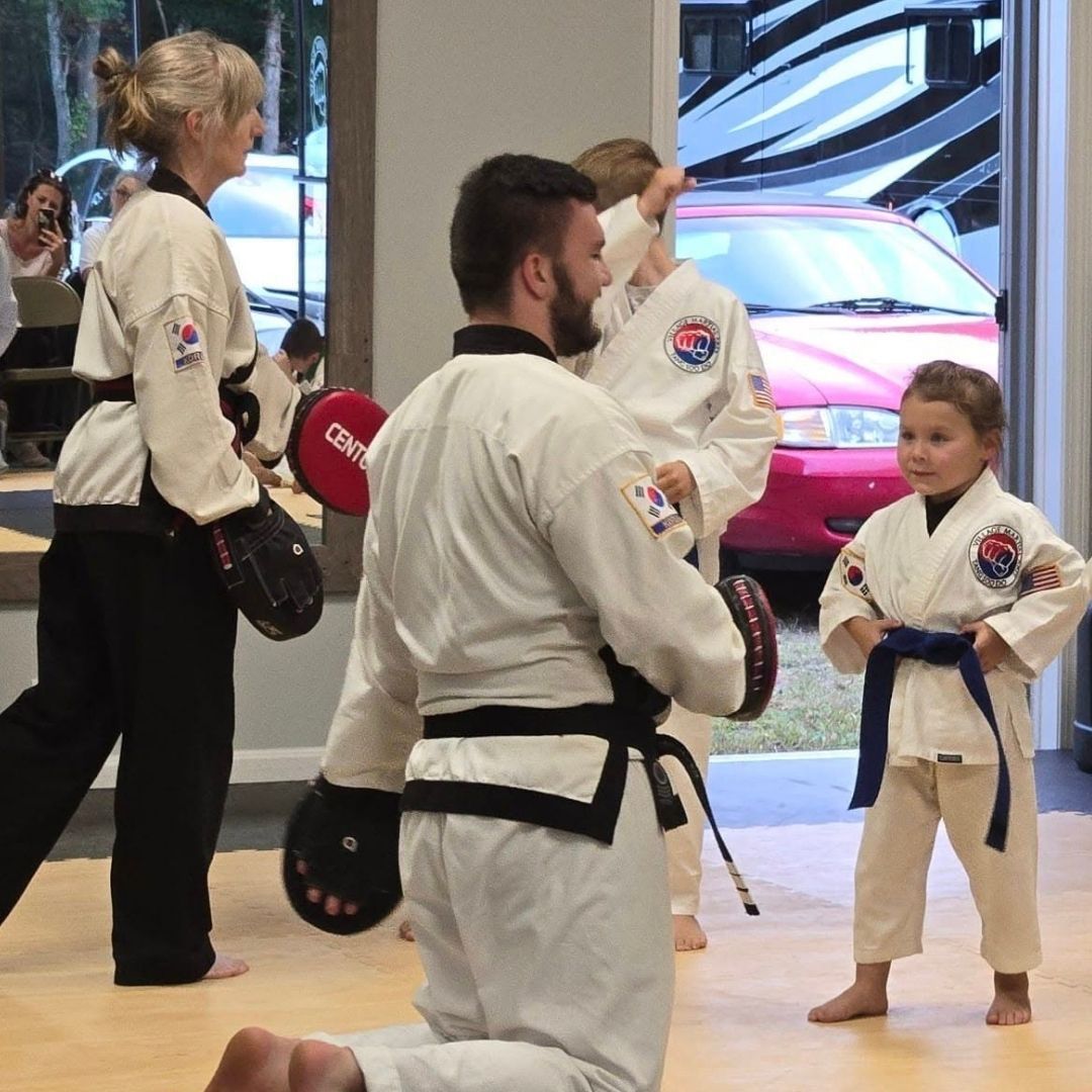 Two children in black and white martial arts uniforms practice a chokehold on a mat. One child smiles.