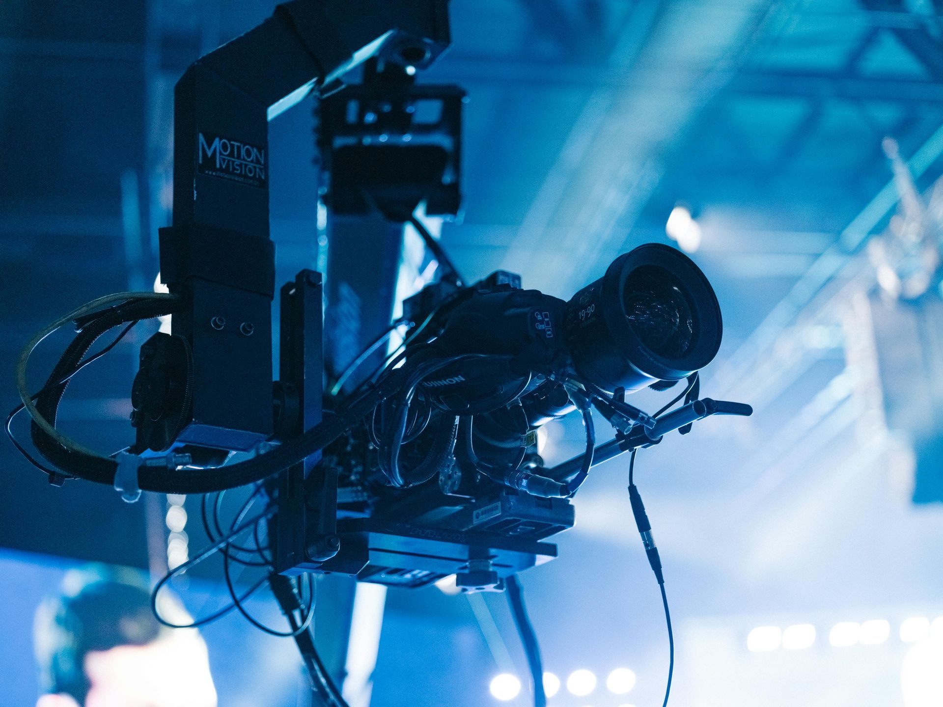 Camera on a crane, aimed toward a bright stage, wires visible, blue lighting.