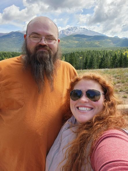 Man with beard and woman with red hair smile in front of a mountain and trees.