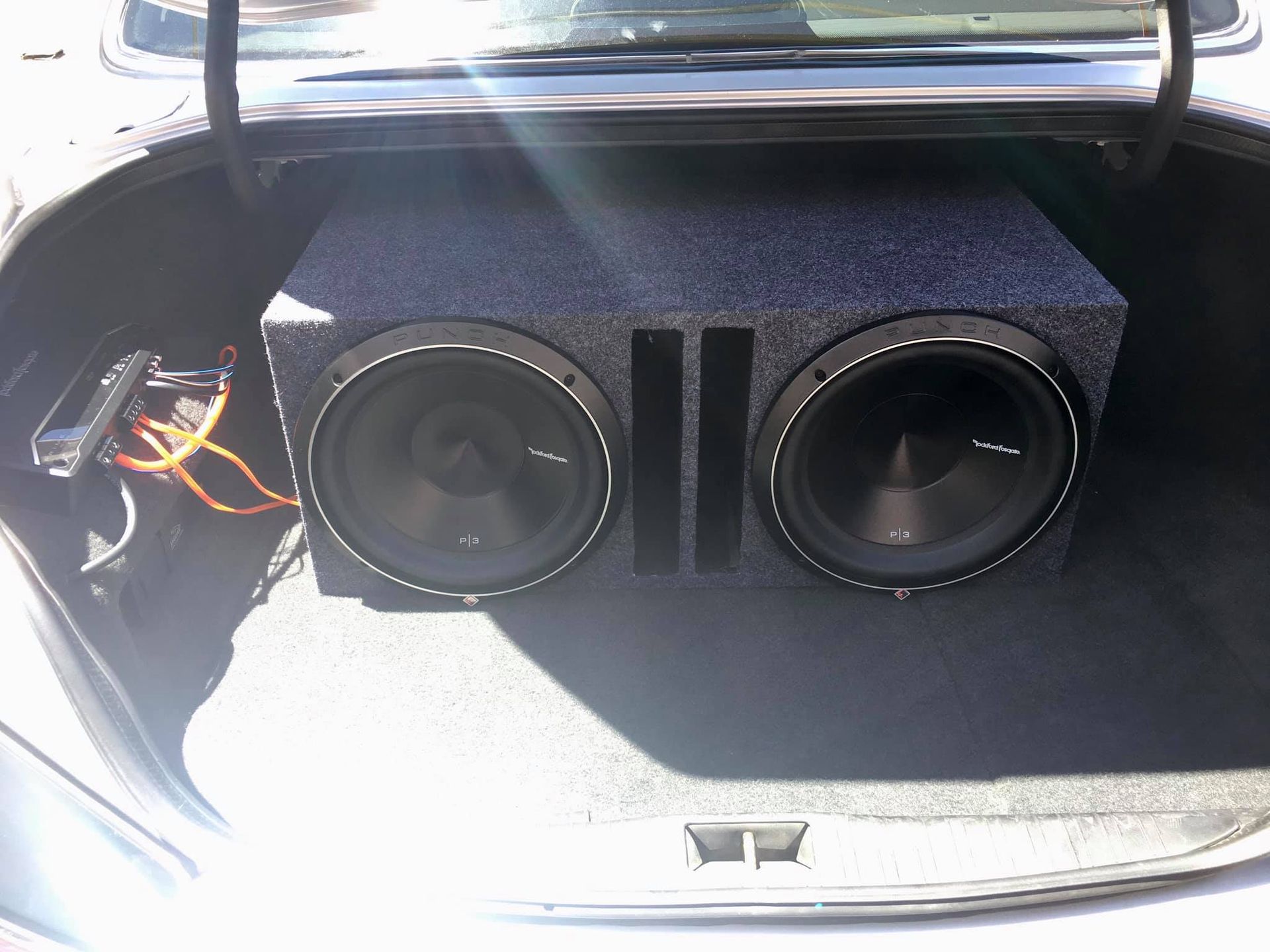 A car trunk with a large dual subwoofer box installed. Black speakers, grey box, and orange wiring are visible.
