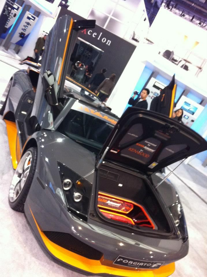 Gray and orange sports car with open doors and trunk at an auto show.