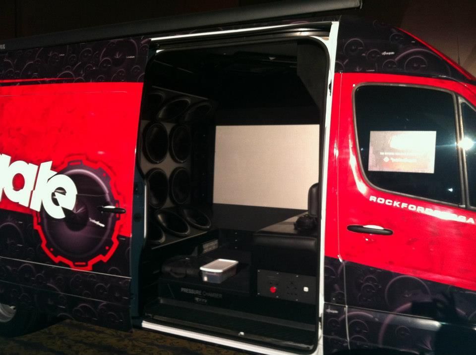 A red and black van with open door reveals a movie theater setup inside, including a screen and large speakers.