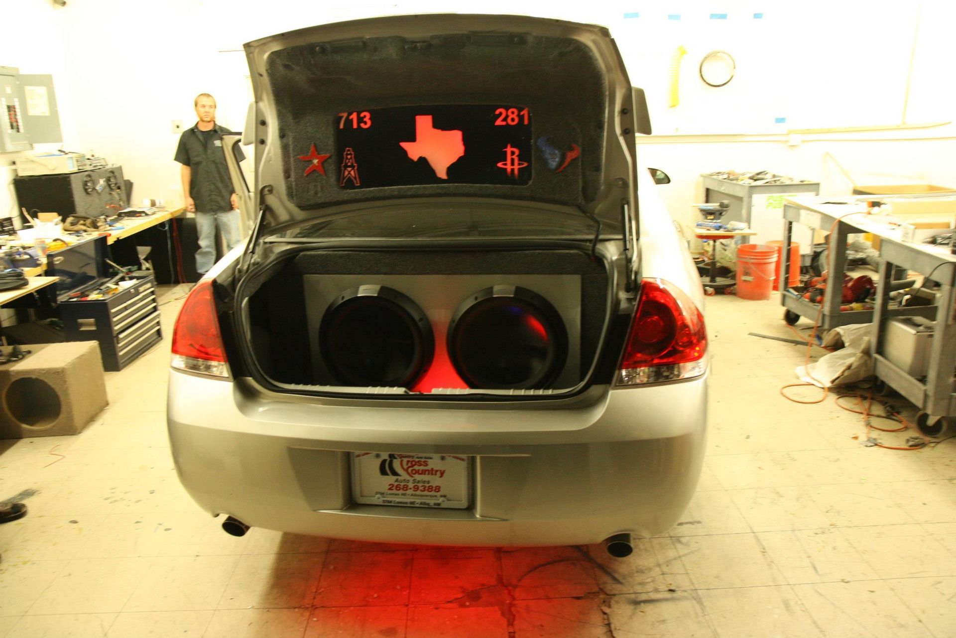 Car trunk with dual subwoofers, Texas-themed display, red underglow. Man stands nearby in a shop.