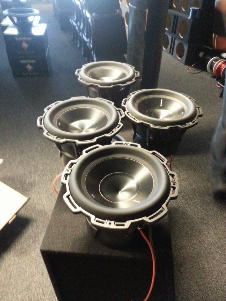 Four black subwoofers with silver trim, wired, on a black box, indoors.