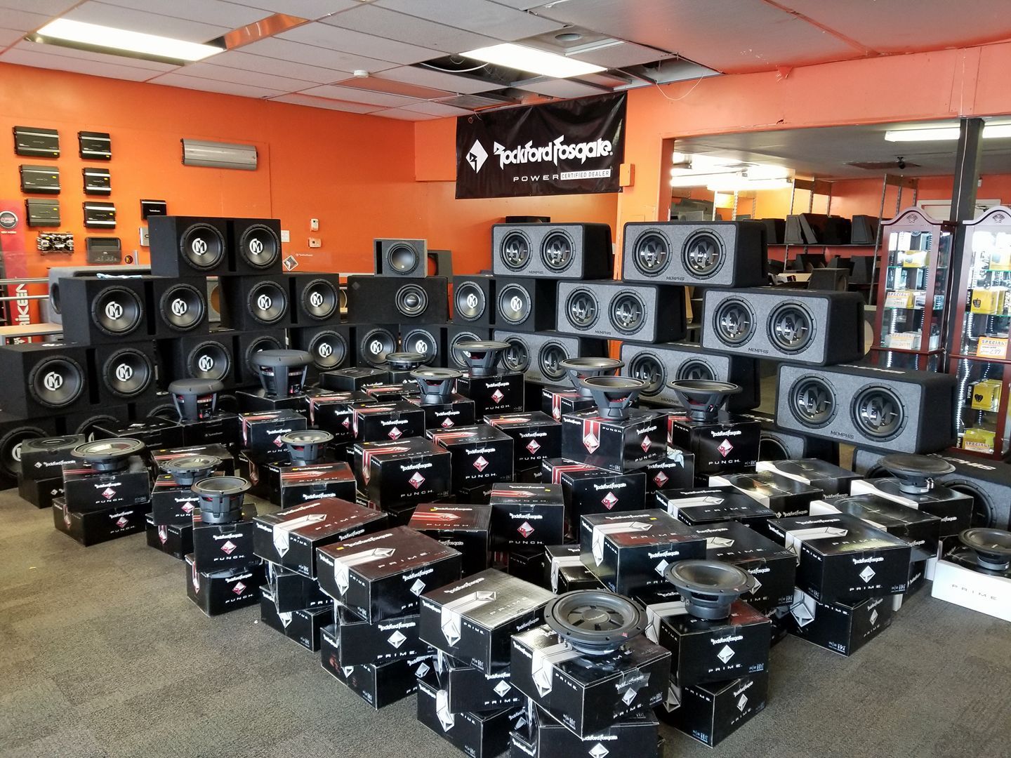 Inside an audio shop, rows of speakers and boxes, mostly black, sit on the floor and shelves. The walls are orange.