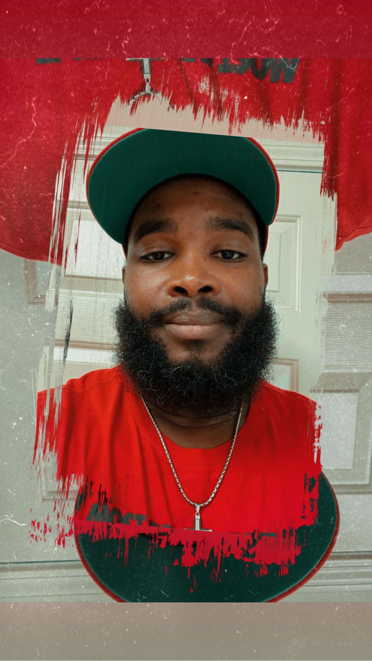 Man with a beard, wearing a red shirt and a green cap, in front of a red background.