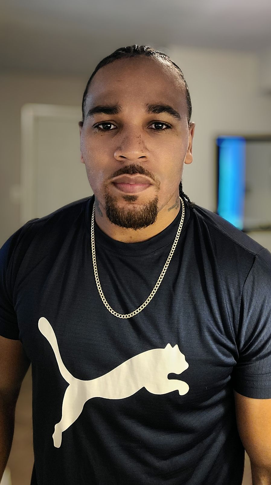 Man with braids wearing a blue Puma shirt and silver chain, looking directly at the camera.