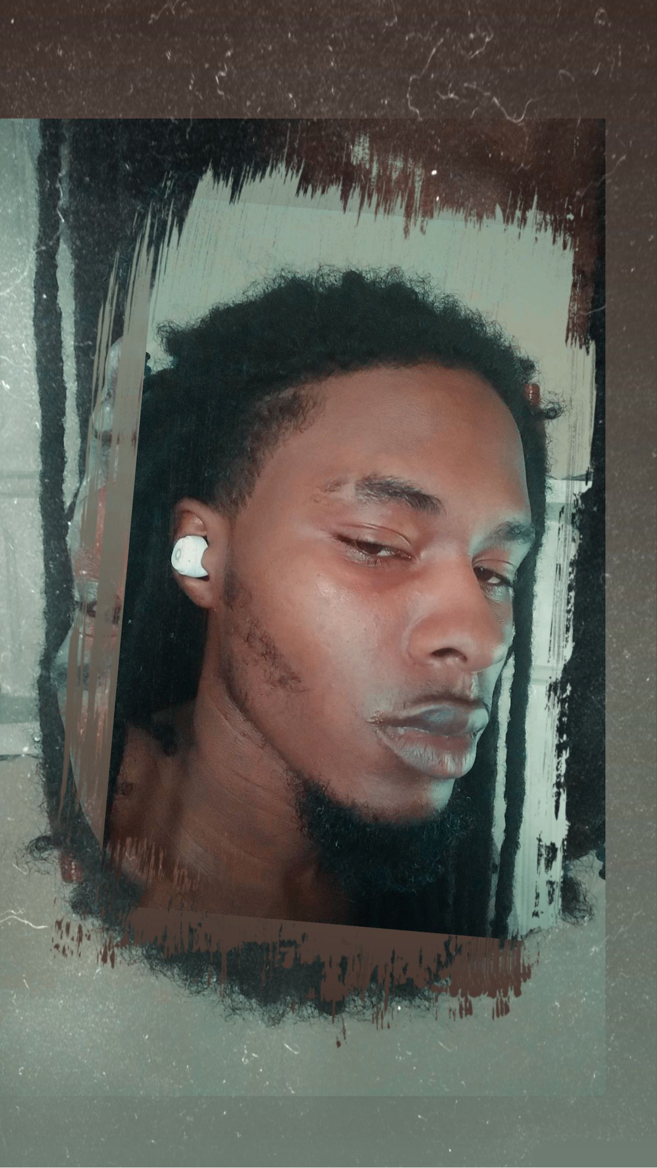 A young Black man with dreadlocks and earbuds gazes downward, set against a distressed, blurred background.