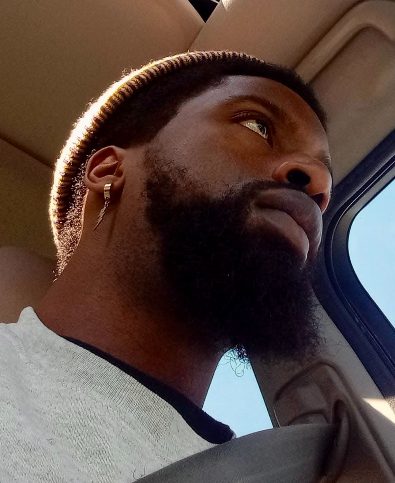 Black man with a beard and earring looks out window with thoughtful expression. Inside a car.