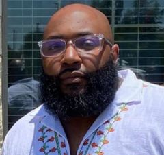 Bald man with beard and glasses, wearing a white shirt with floral design, looking at camera.