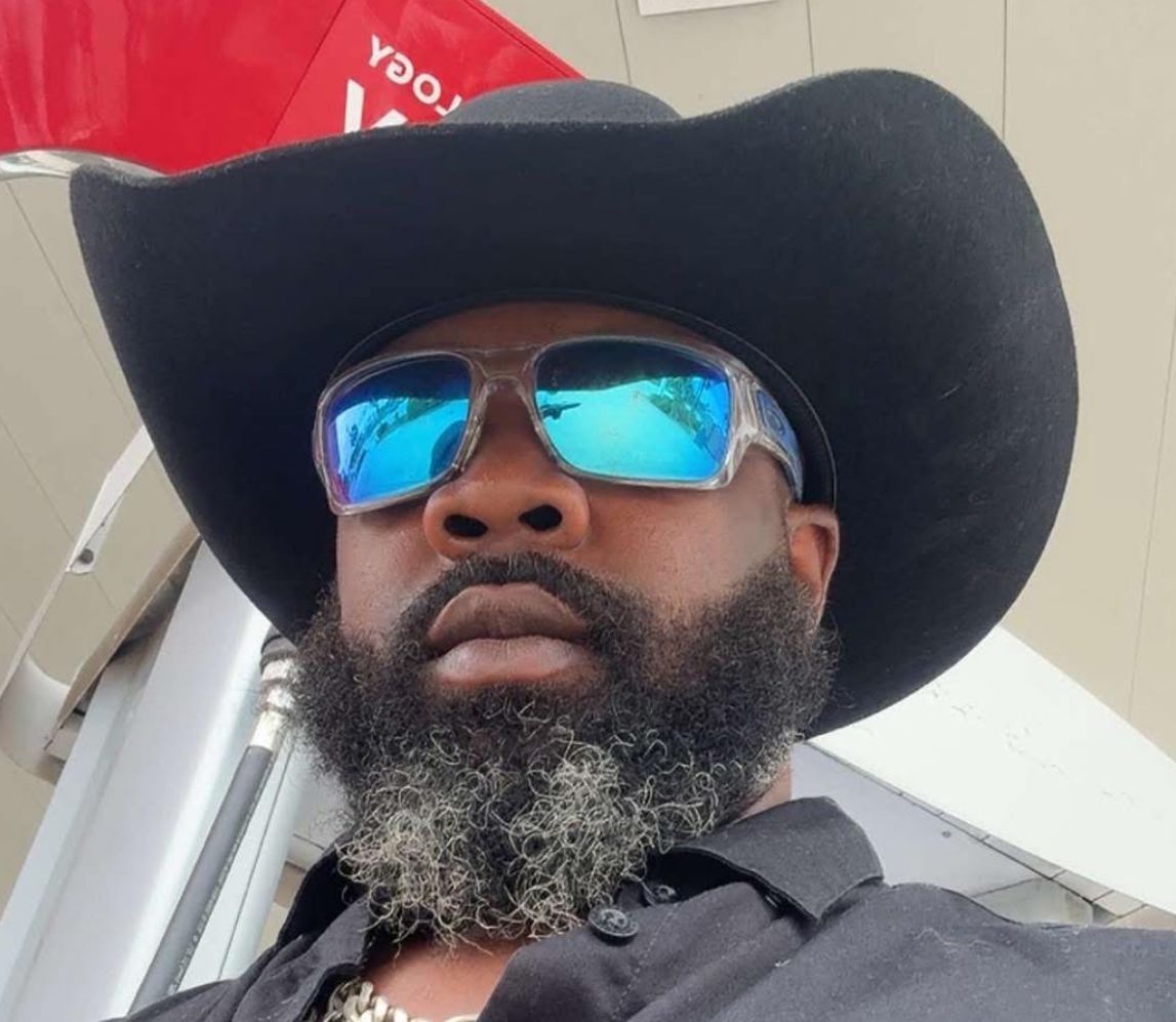 Man with sunglasses and cowboy hat, outdoors. He has a full beard and is looking at the camera.