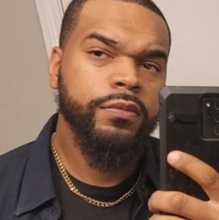 A Black man with a beard, holding a phone up to take a selfie. Wearing a gold chain and dark clothing.