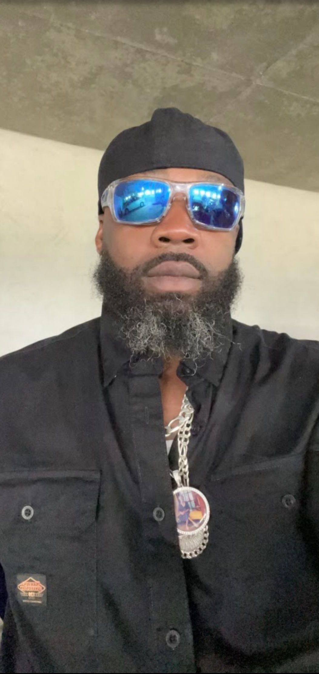 A Black man wearing sunglasses and a black cap, with a black shirt and a silver chain.