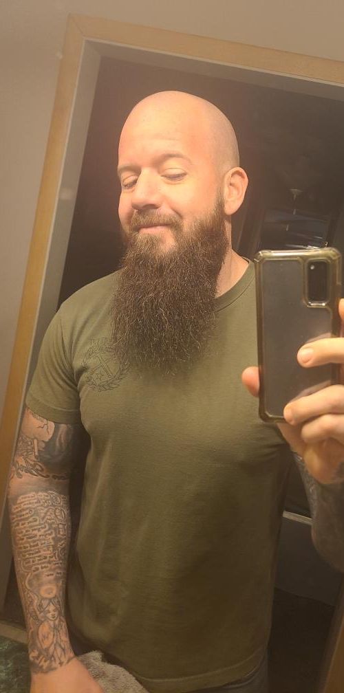 Bald man with a long beard wearing a green shirt, taking a selfie in a mirror. He has tattoos on his arm.