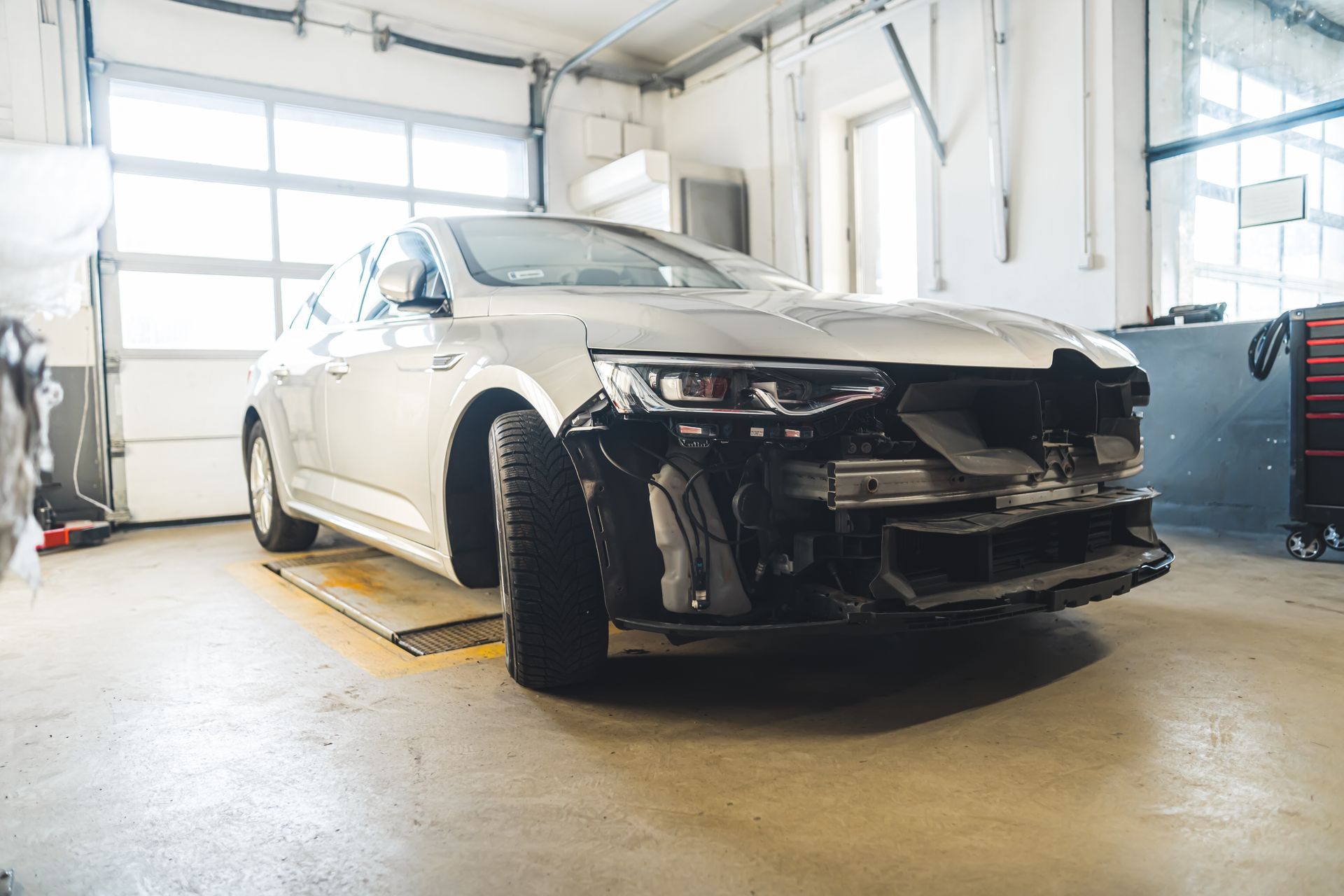 A silver car with a damaged front end is parked in a garage.