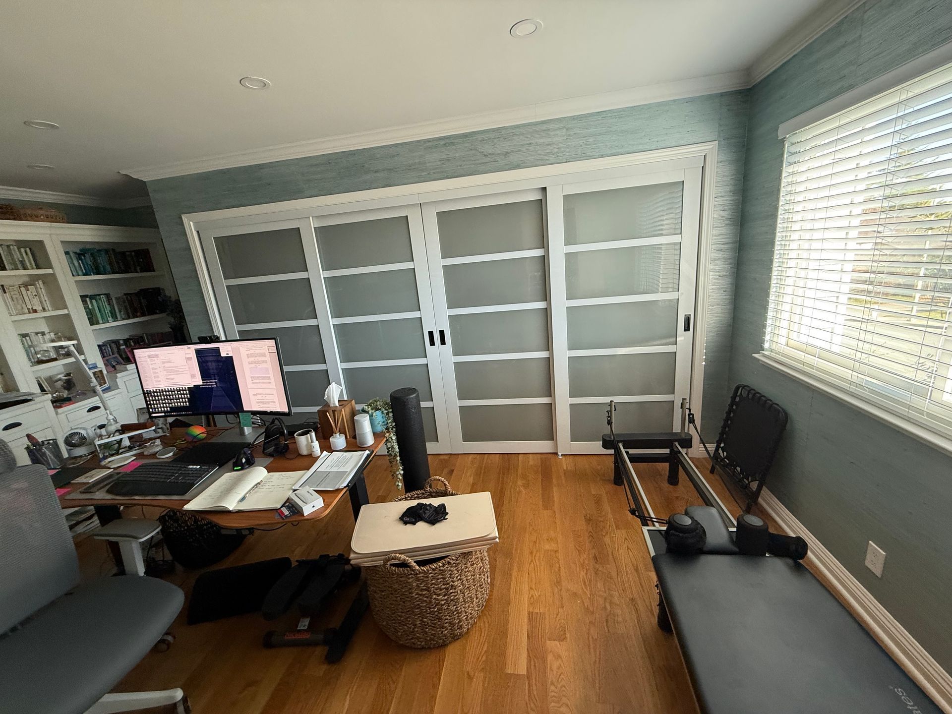 Home office with frosted glass sliding doors, a pilates reformer, and a desk with a computer. Light blue walls and wood flooring.