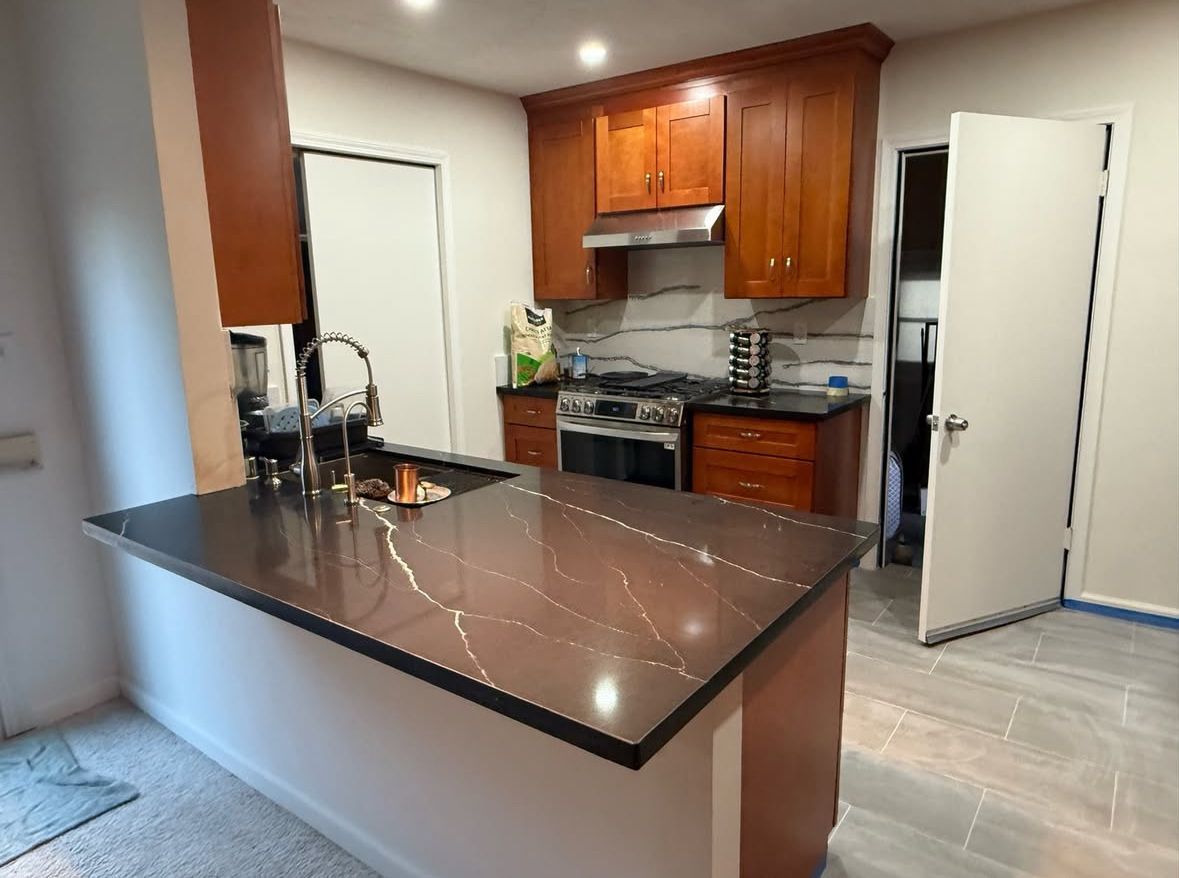 A modern kitchen with brown cabinets, a dark countertop, and an island. A stainless steel stove and sink are visible.