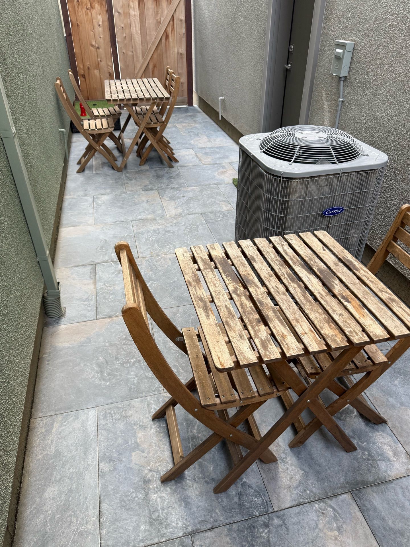 Two small wooden tables with chairs on a stone patio in a narrow outdoor space. An air conditioning unit is present.