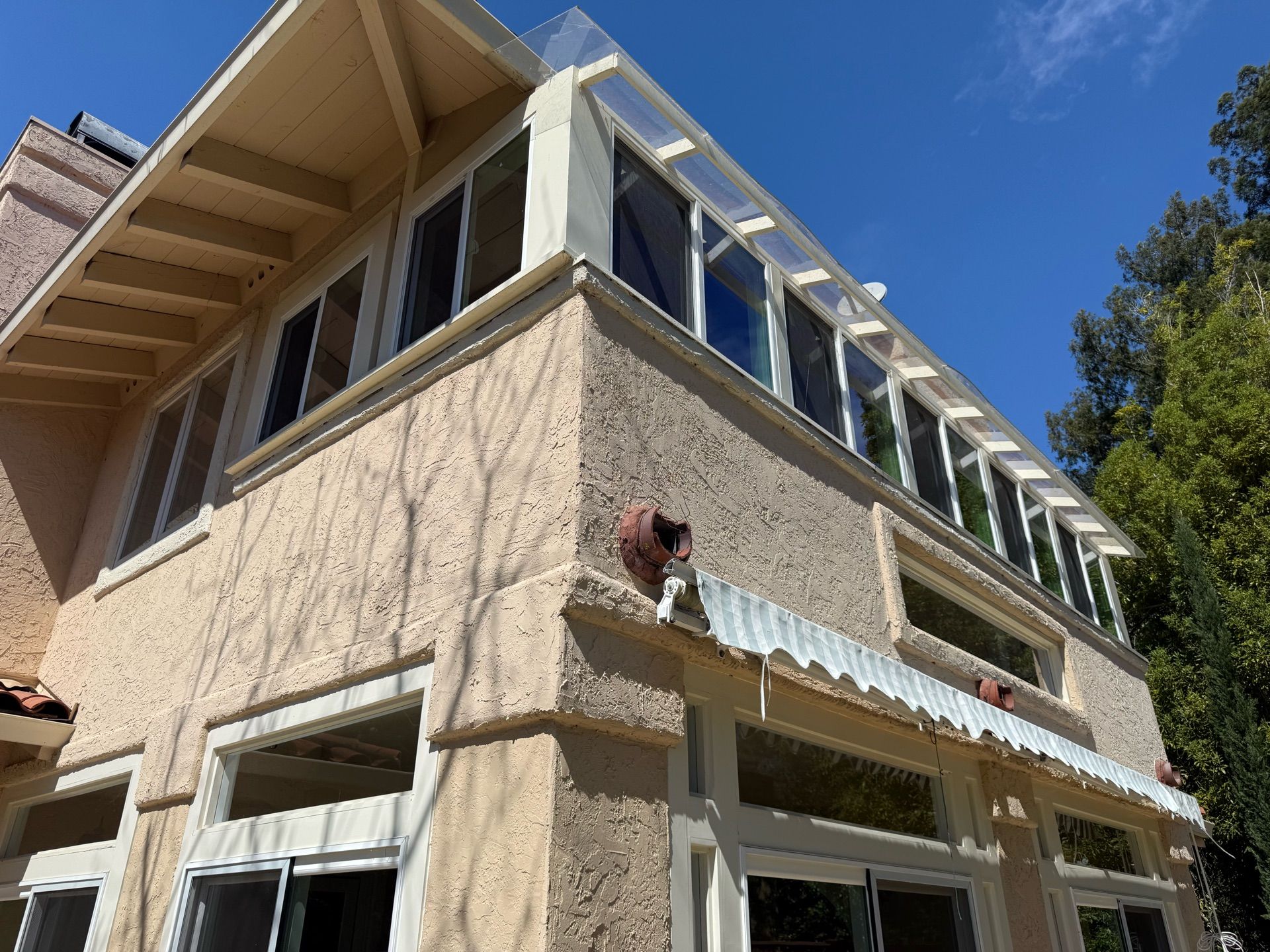 Two-story stucco building with many windows. A clear roof extends from the upper level, with green trees and blue sky visible.